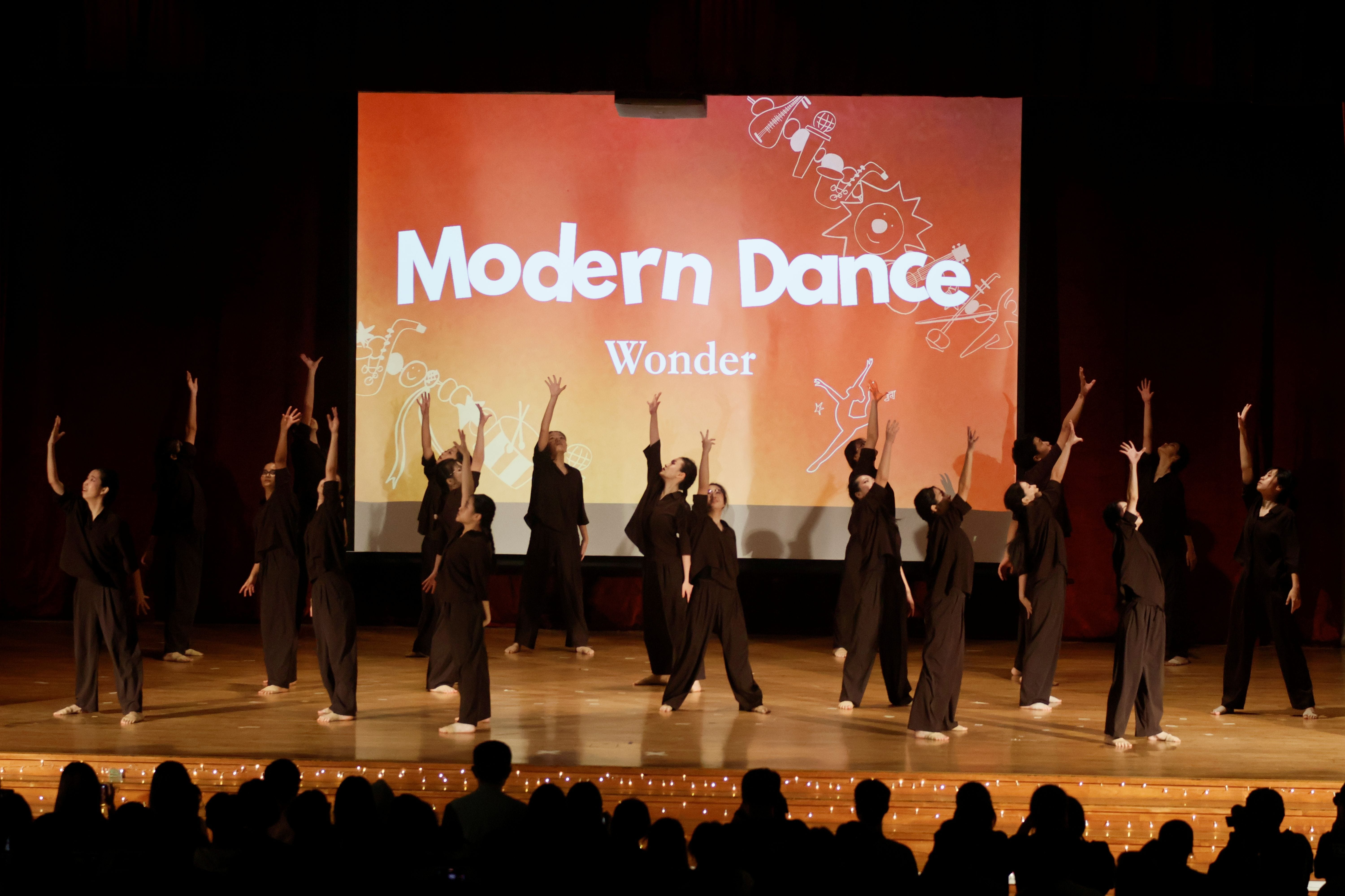 A large group of dancers in black costumes perform "Wonder", arms stretched upward in unison on a warmly lit stage.